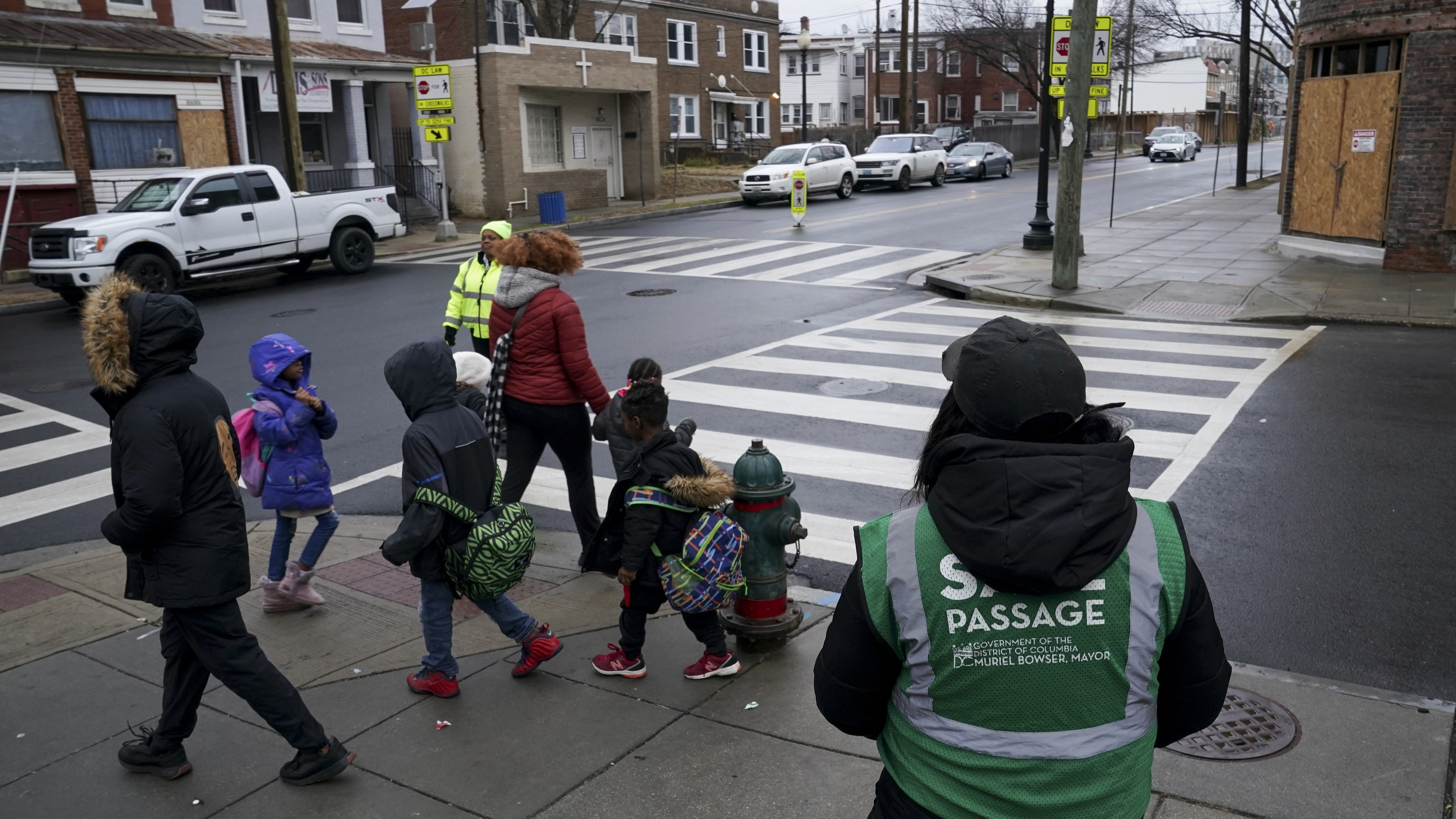 A group of elementary school children wearing winter coats and backpacks stand on a street corner outside an elementary school, waiting for crossing guards