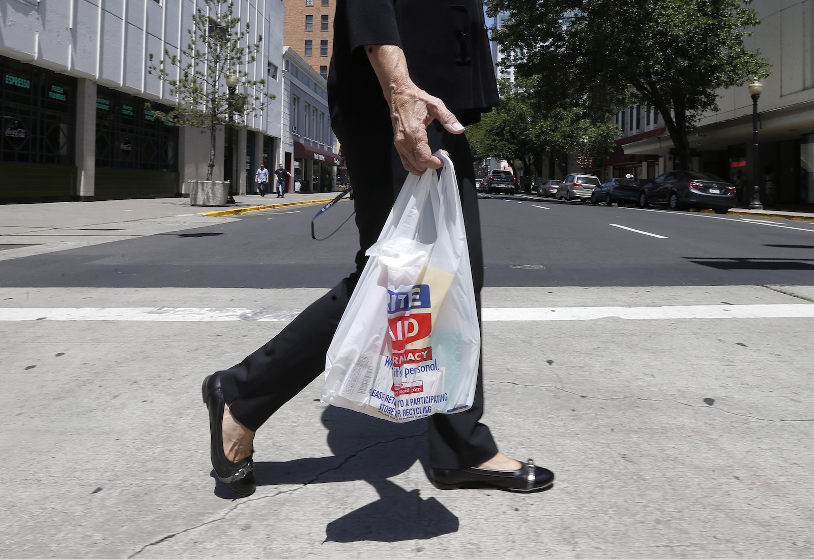 Cropped view of a shopper dressed in black, walking while holding a Rite Aid brand plastic bag.