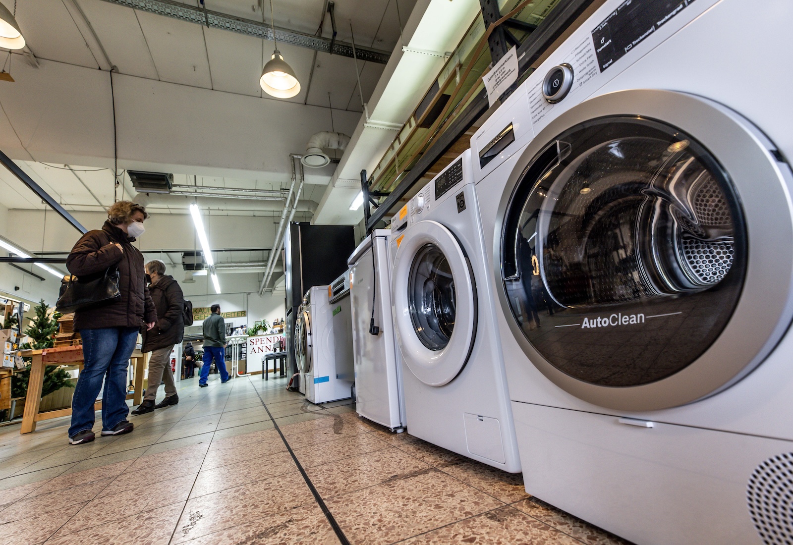 White washing machines are lined up on a shop floor, with shoppers looking at them.