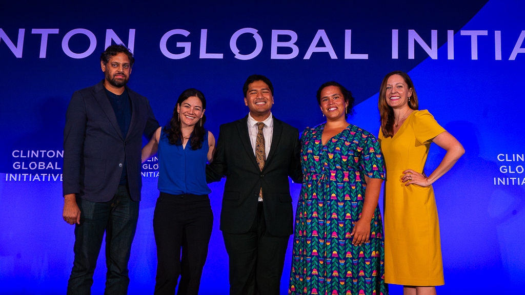 Five people stand on a stage smiling in front of a Clinton Global Initiative backdrop