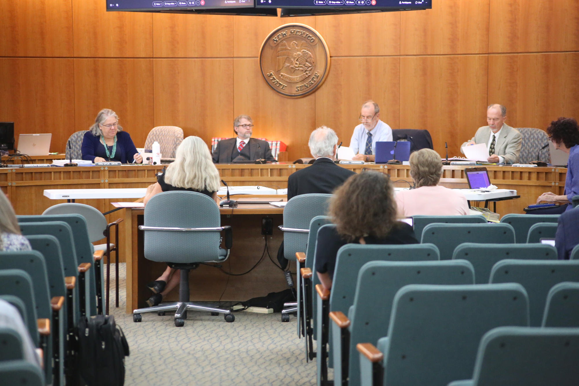 A wood paneled meeting room with blue chairs contains people in front speaking to members of the audience