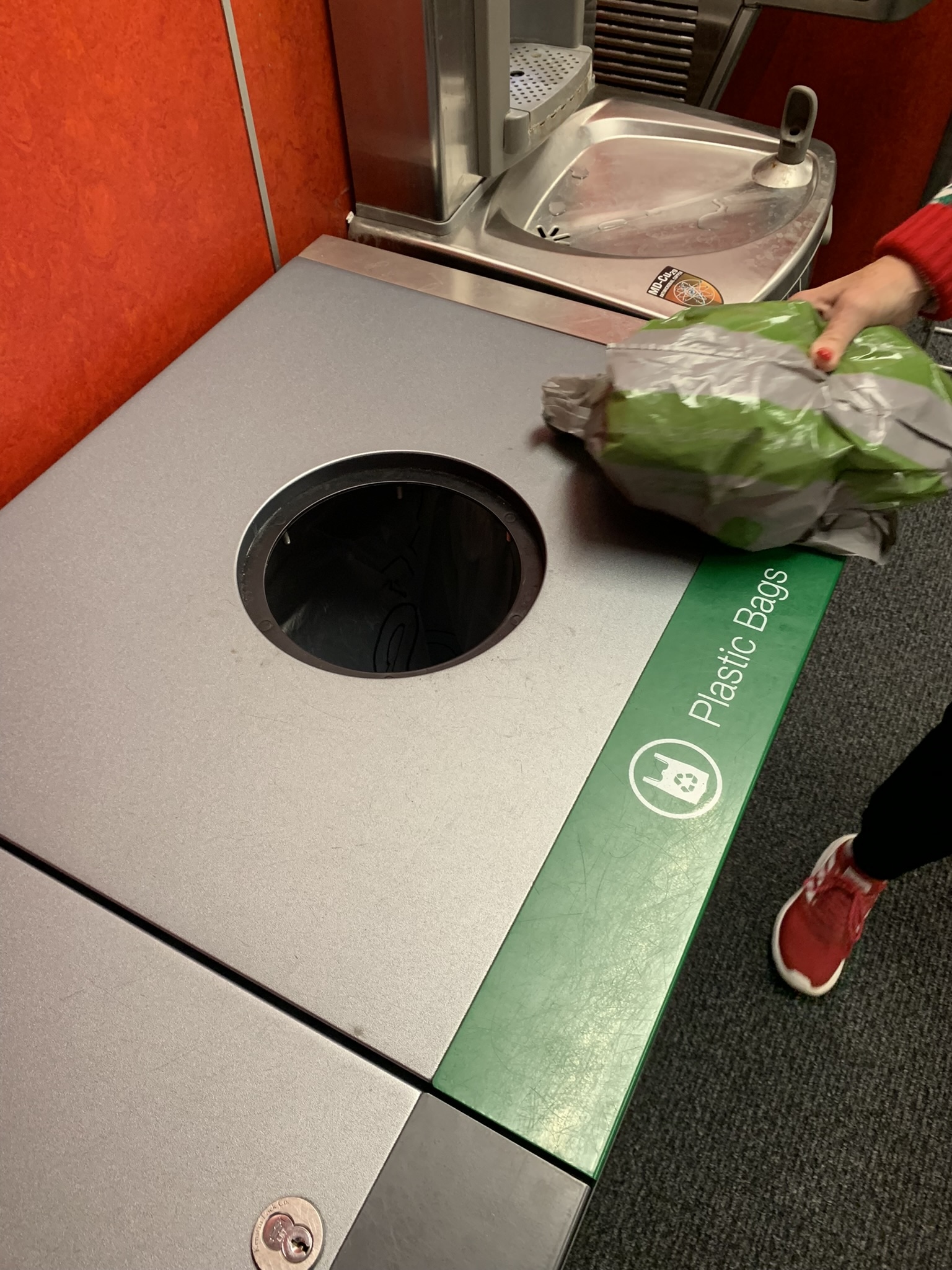 A circular receptacle labeled "plastic bags," with a woman's hand reaching toward it to deposit a plastic bag for recycling.