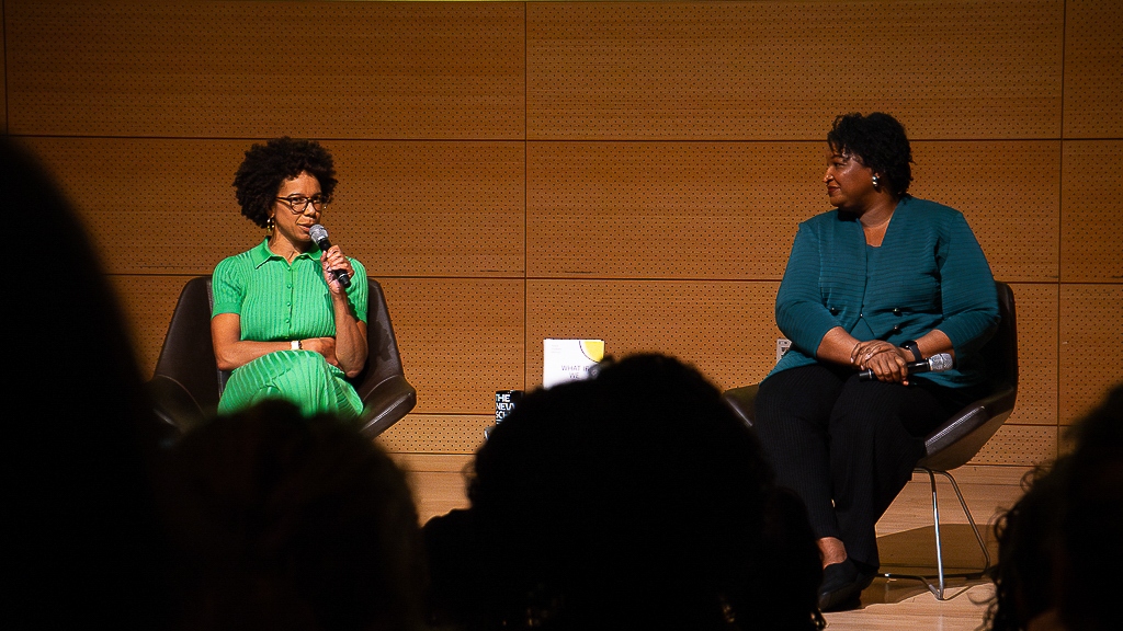 A woman in a green dress sits on a stage next to a woman in a blue shirt talking on microphones