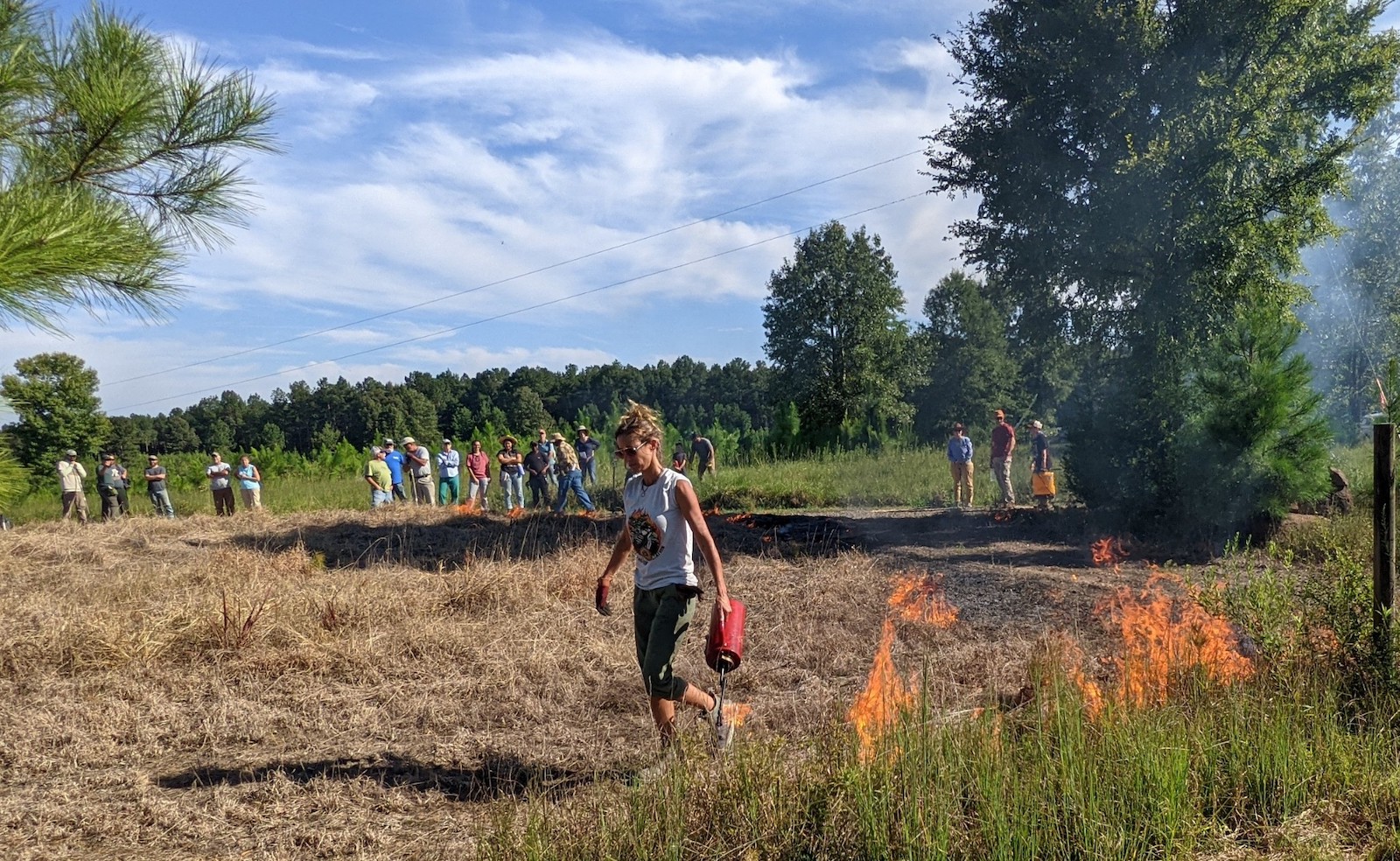 A woman holds a small canister setting controlled fires while walking through a grassy field