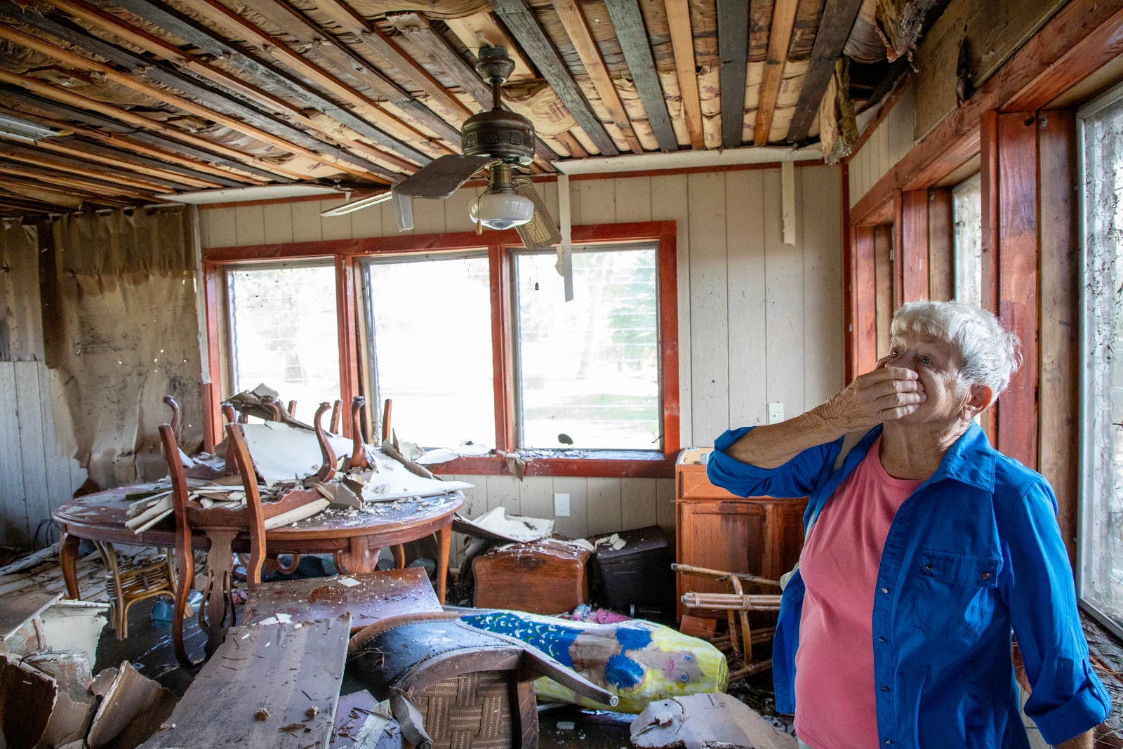 A woman with white hair holds her hand over her mouth and nose while standing in the aftermath of her flooded home