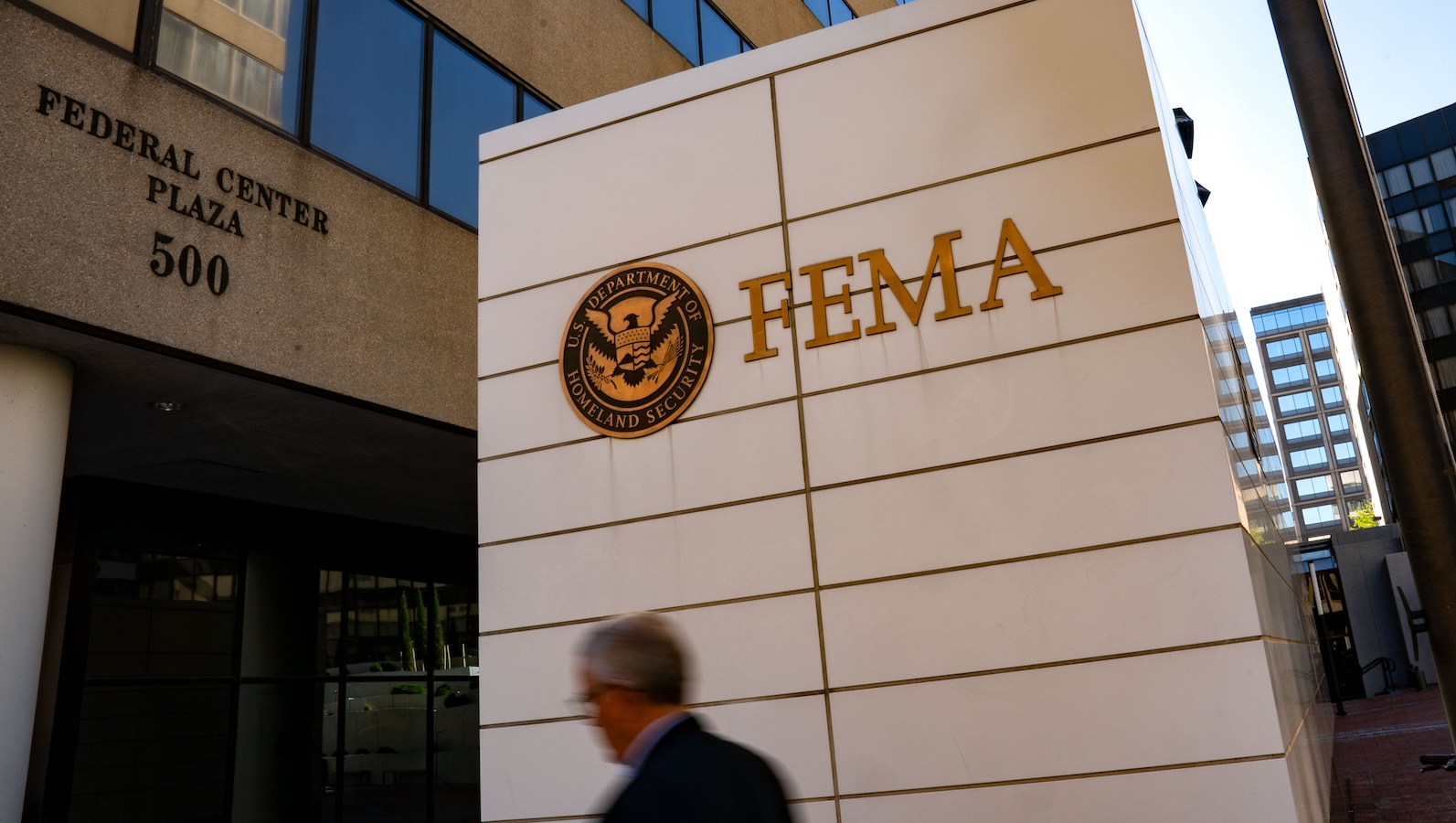 A shot of a man from the shoulders up walking past the Federal Emergency Management Agency headquarters in Washington, DC. The FEMA logo and address can be seen on the exterior of the building