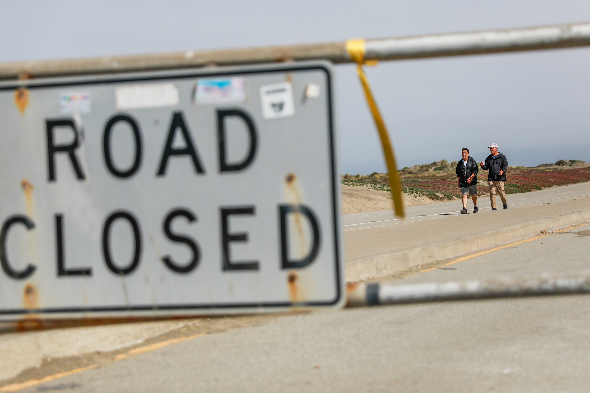 A large sign that says 'road closed' blocking a highway near a beach. Beyond, two people walk on the road