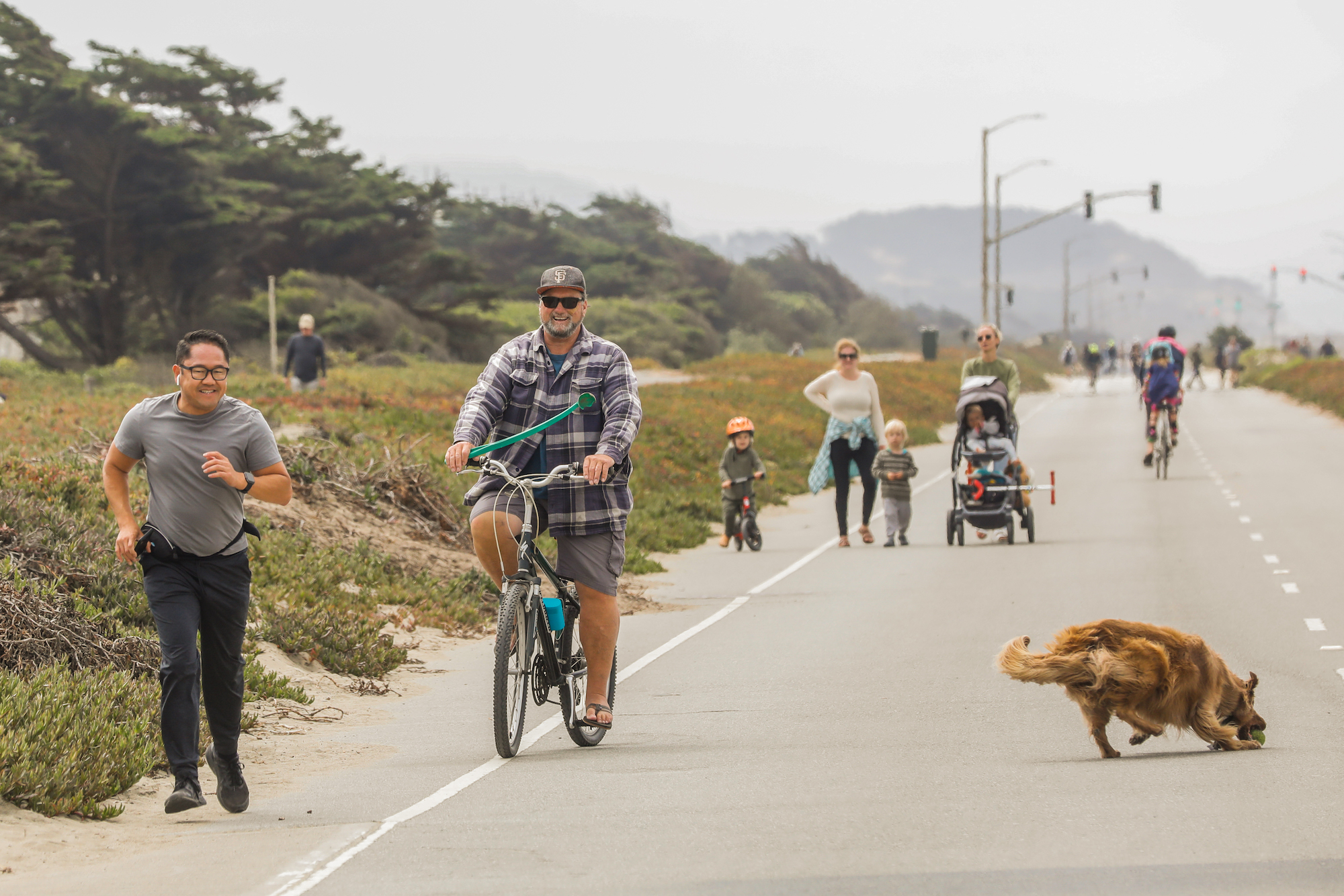 bikers and joggers smile as they move along a coastal highway. In the foreground, a dog sniffs the road. In the background, people jog with strollers.