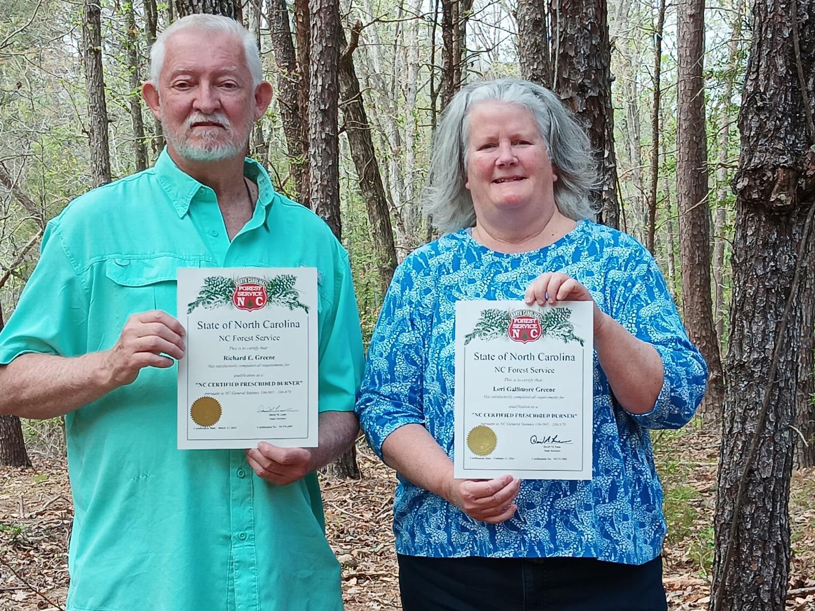 An elderly man and woman hold certificates standing in front of a forest