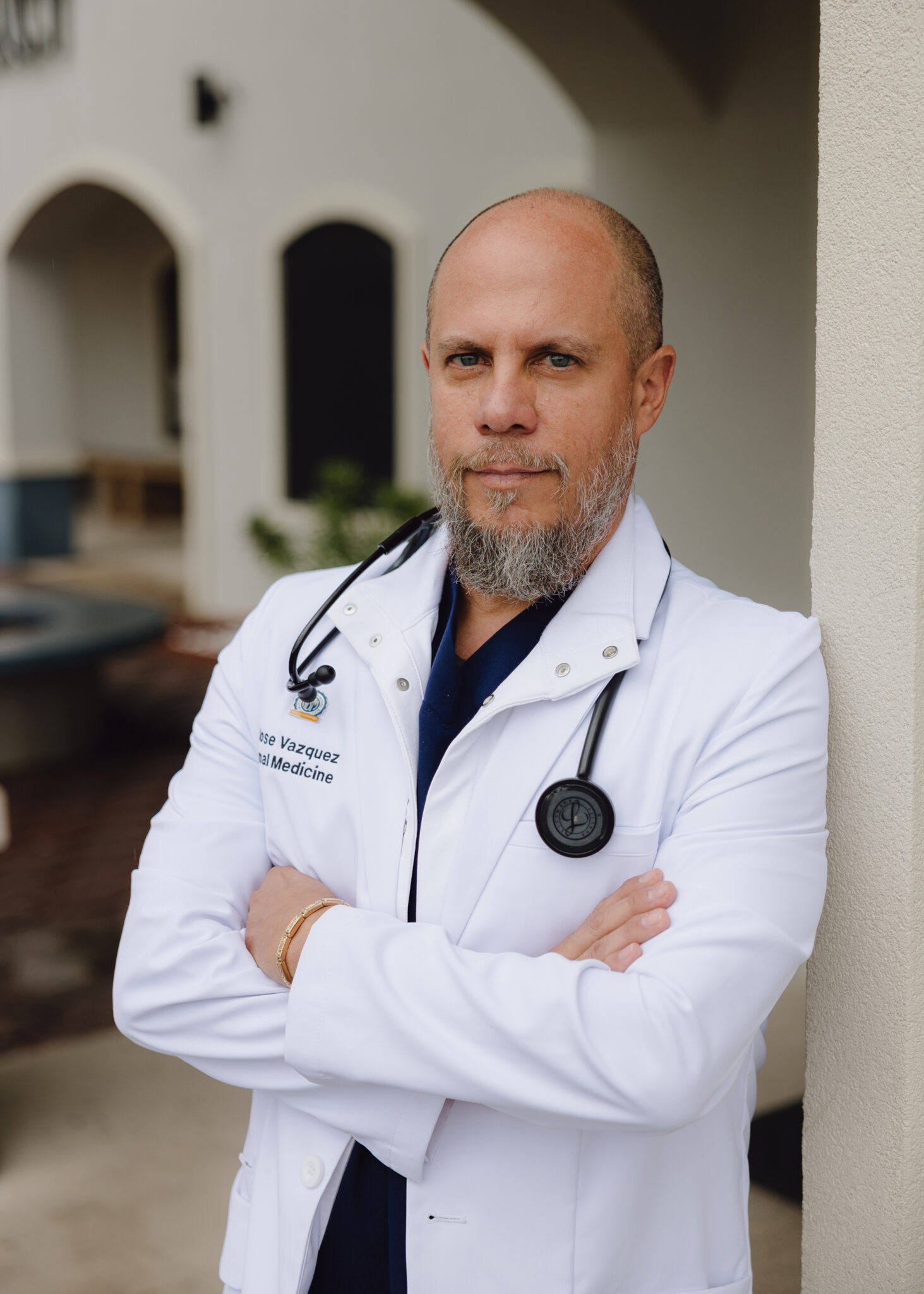 A bald man with a gray beard wears a doctor coat and stethescope