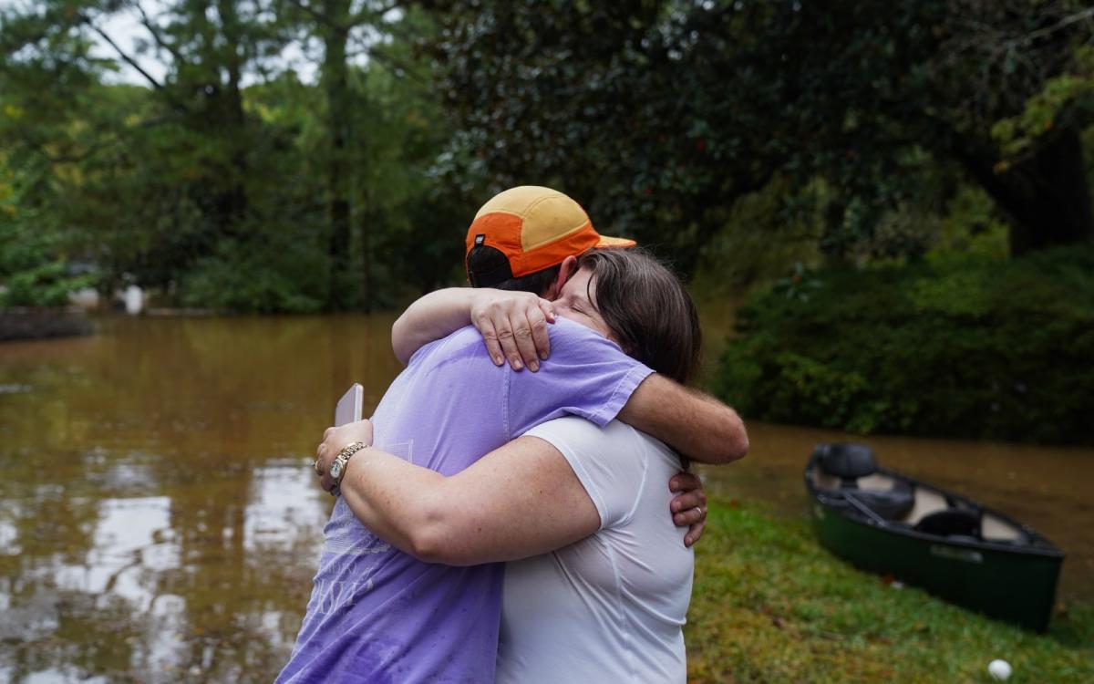 Two people hug tightly in front of a flooded field, with a small black canoe sitting in the background