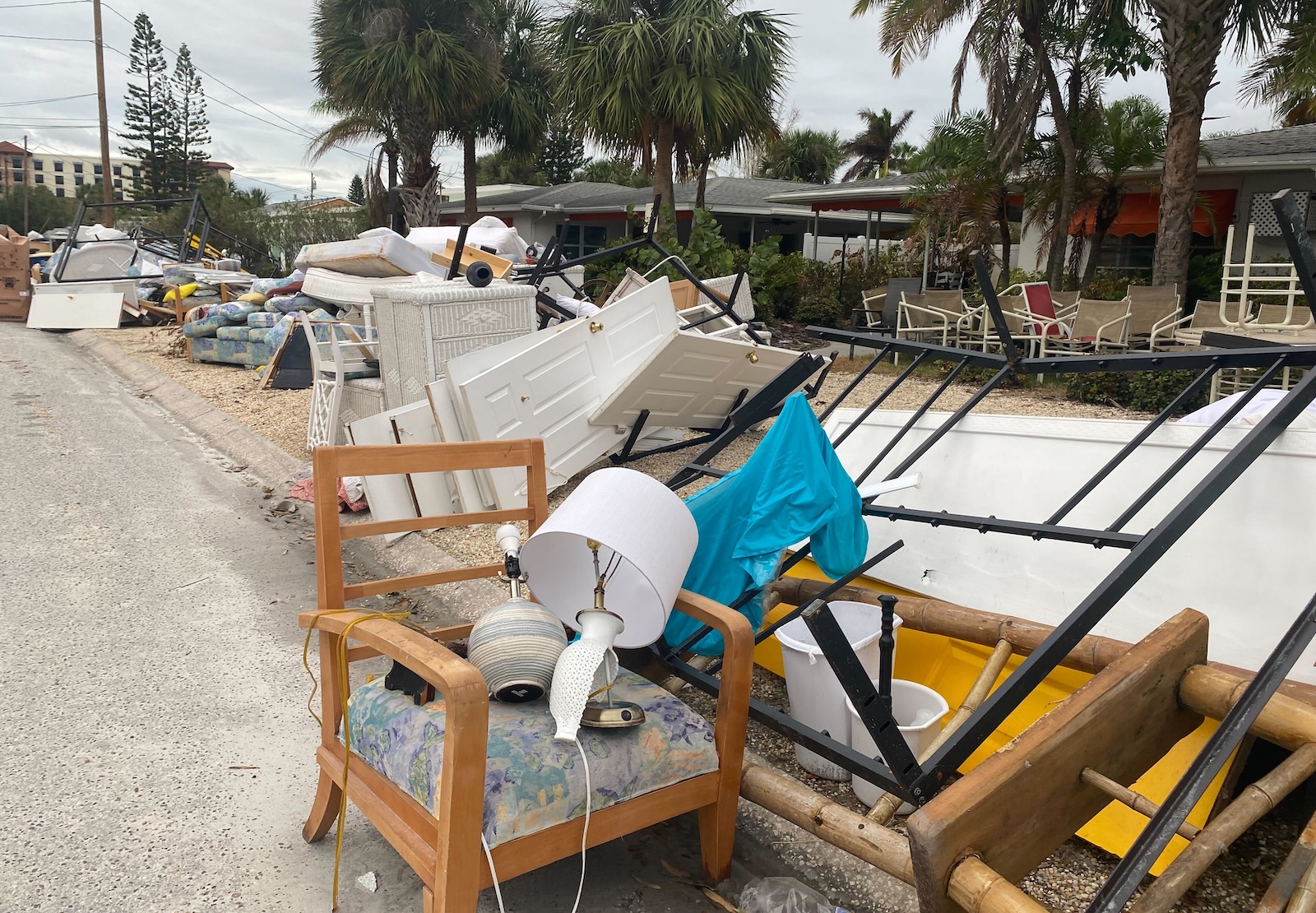 A chair, lamp, and other debris are strewn on the street outside a row of houses, with palm trees in the background.