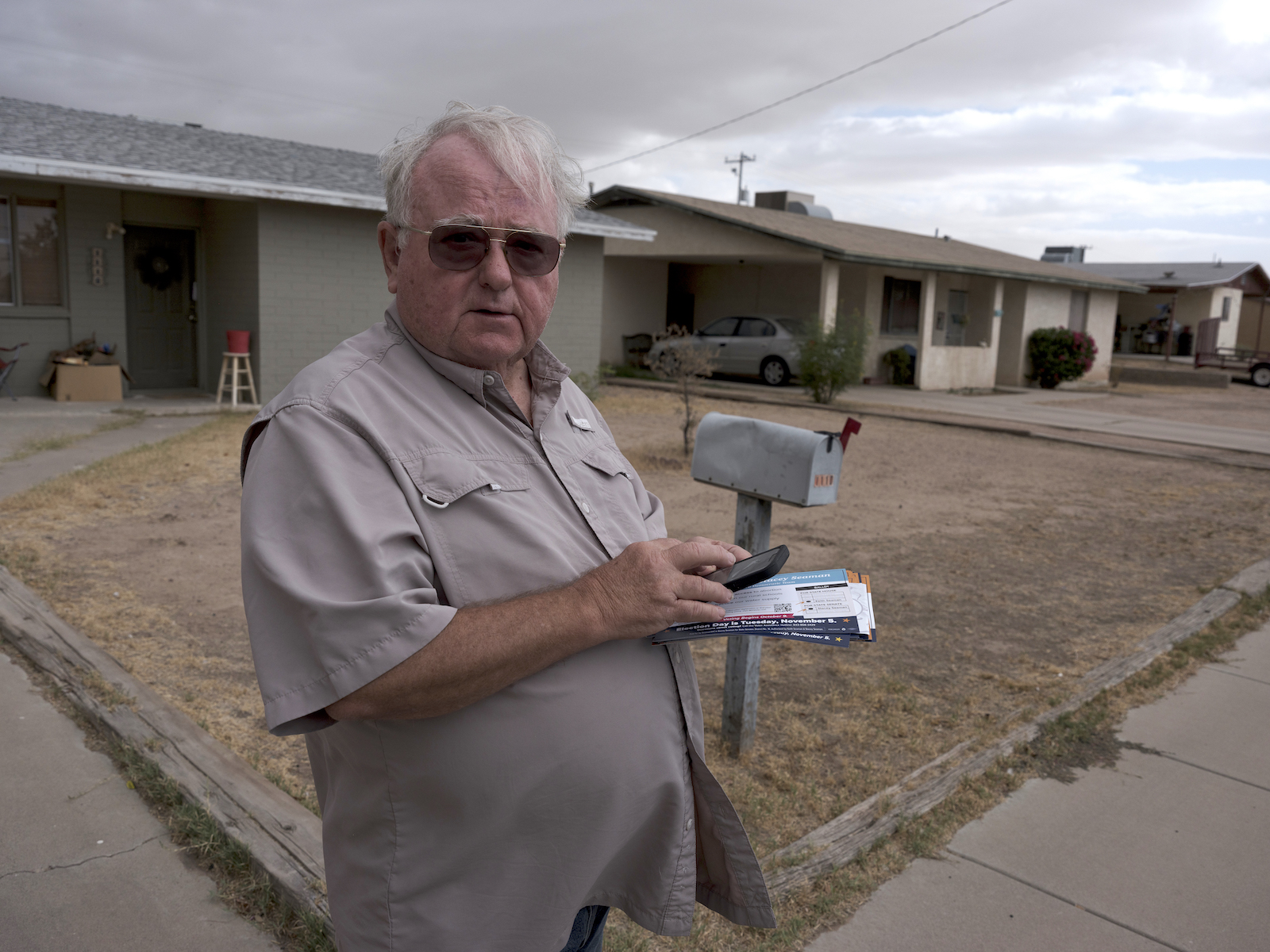 A man in a button-up shirt wears aviator sunglasses and looks up from holding flyers near a house