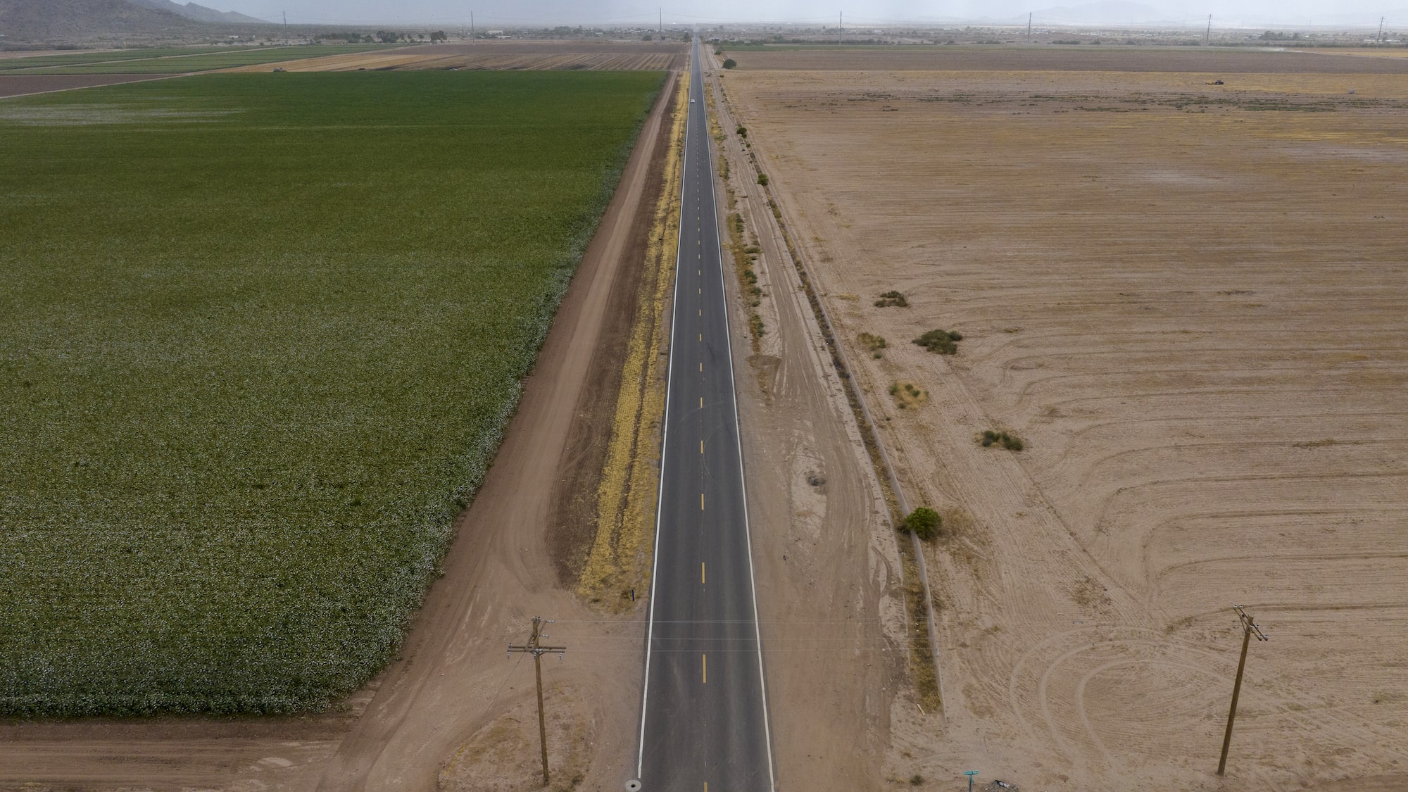 A drone shot of a green field on the left and a dry field on the right separated by a road