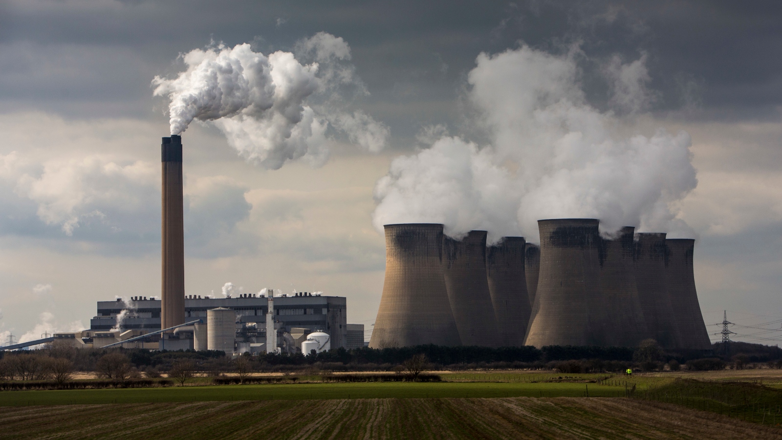 Sitting in a green field, a power plant with smoke blocks blows white smoke into the air