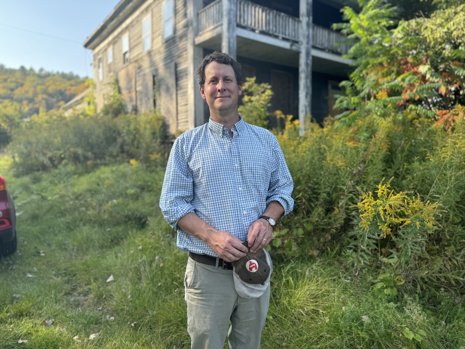 A man in a button up shirt stands in front of a dilapidated house with peeling paint