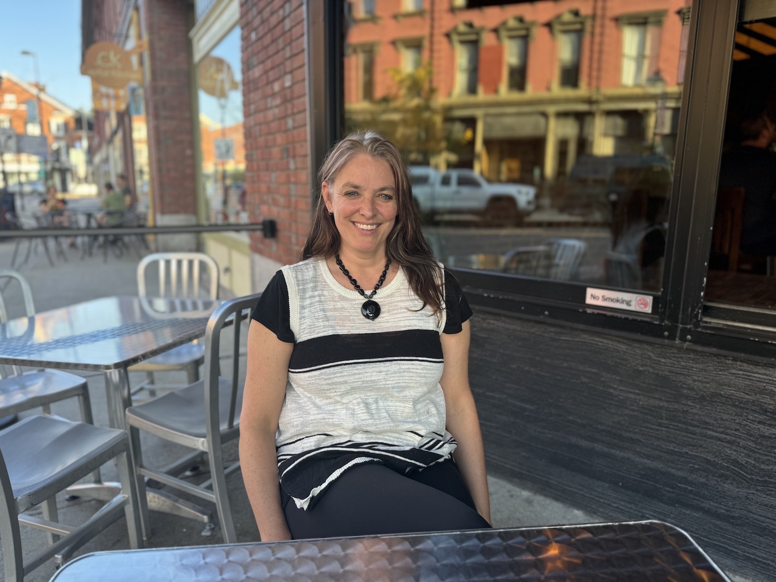 A woman sits at a cafe table smiling near brick buildings