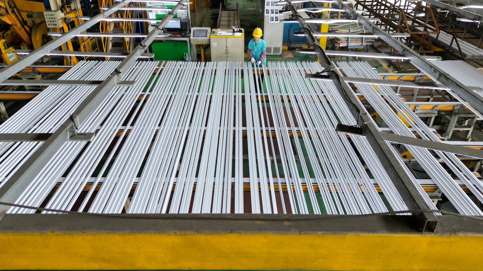 Overhead view of several silver metal strips sitting on top of equipment, with a person wearing a green shirt and a yellow hard hat in the background