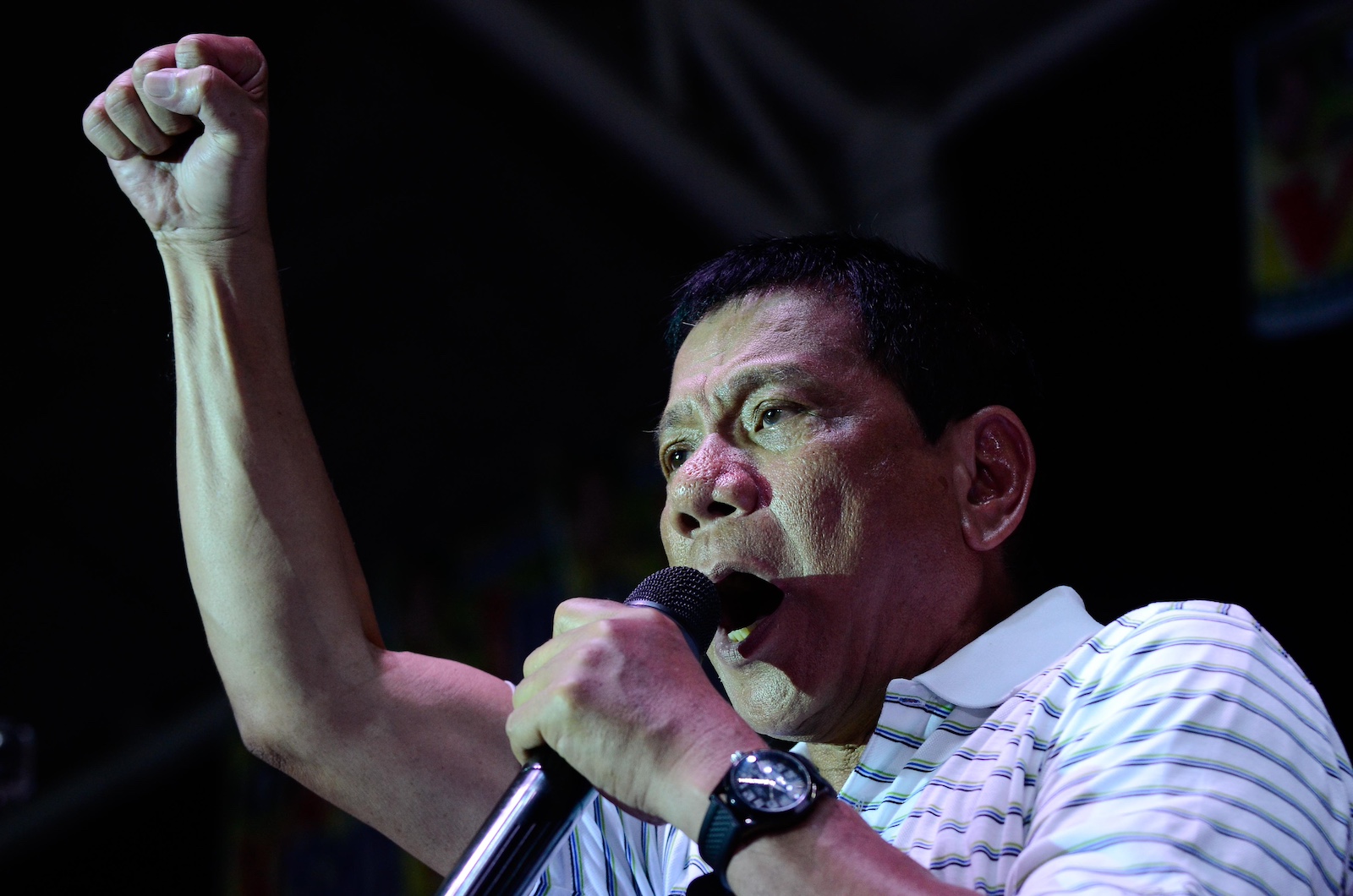 A man wearing a striped black-and-white polo shirt, seen in 3/4 profile, raising his right fist in the air, against a black background