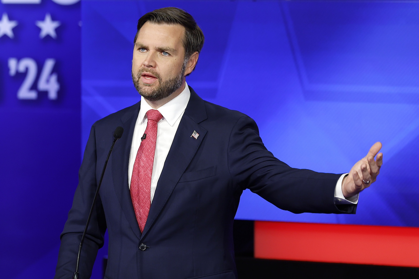 A man with brown hair, a beard and blue eyes, in a suit with a red tie, stands in front of a blue screen with his left arm outstretched