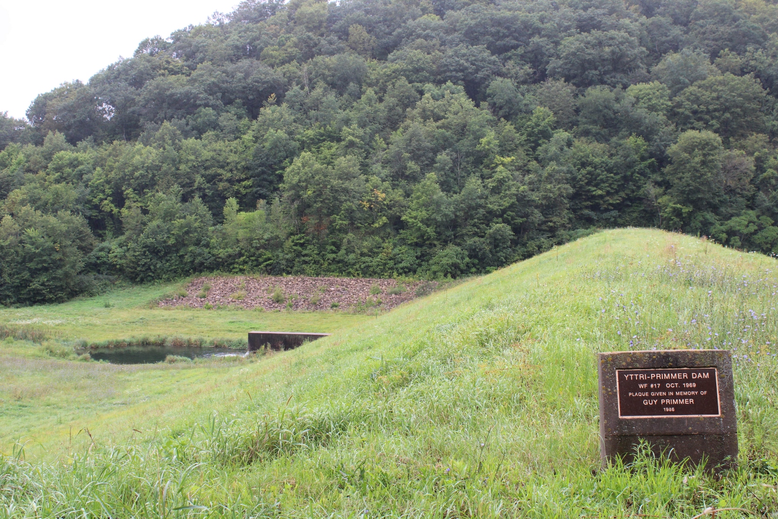 A green hill with a plaque dedicated to the Yttri Dam, which is seen in the background