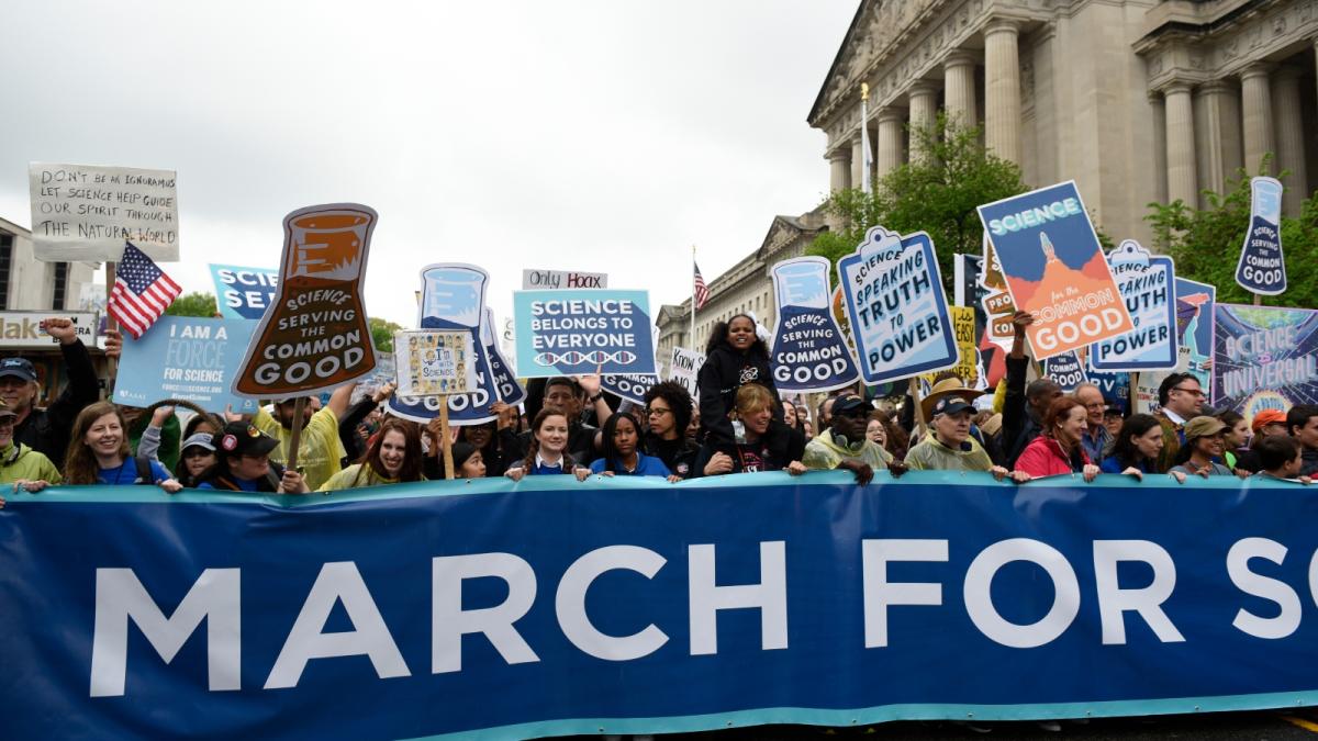 A group of people stand with signs behind a March for Science banner on a street in DC