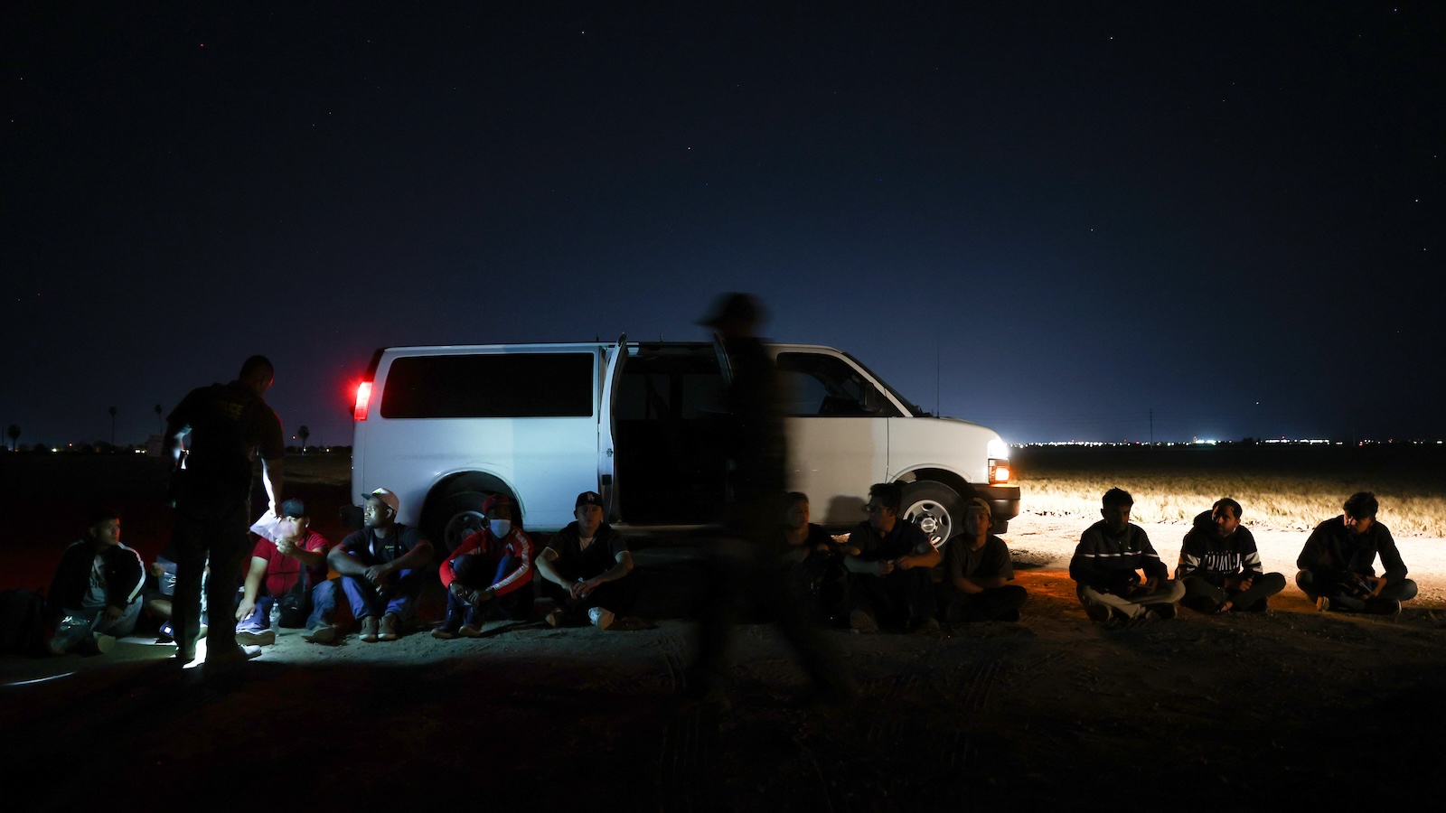 A shot of men sitting in a row in front of an unmarked white van in the middle of the desert. Two border patrol officers can be seen in front of the men, one of them shining a light on the men. The photo is taken during the nighttime, and the only other light comes from the van's headlights.