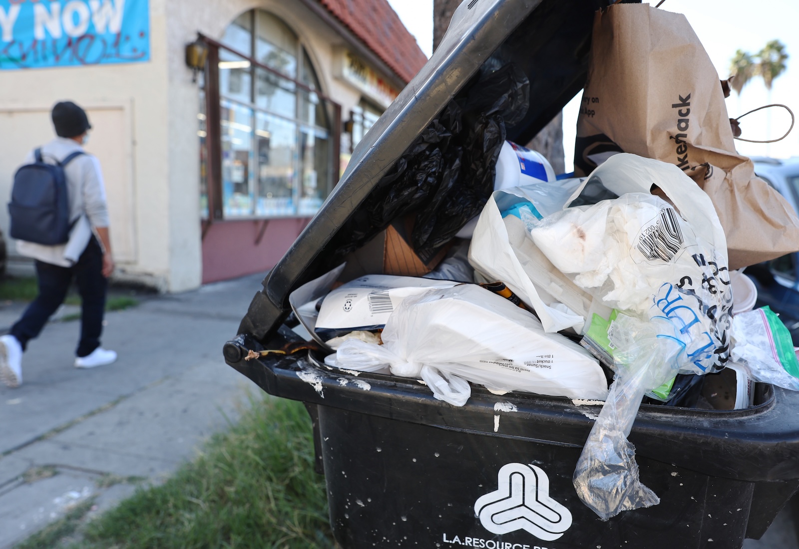 An overflowing trash can with plastic and other trash. A person is walking in the background.