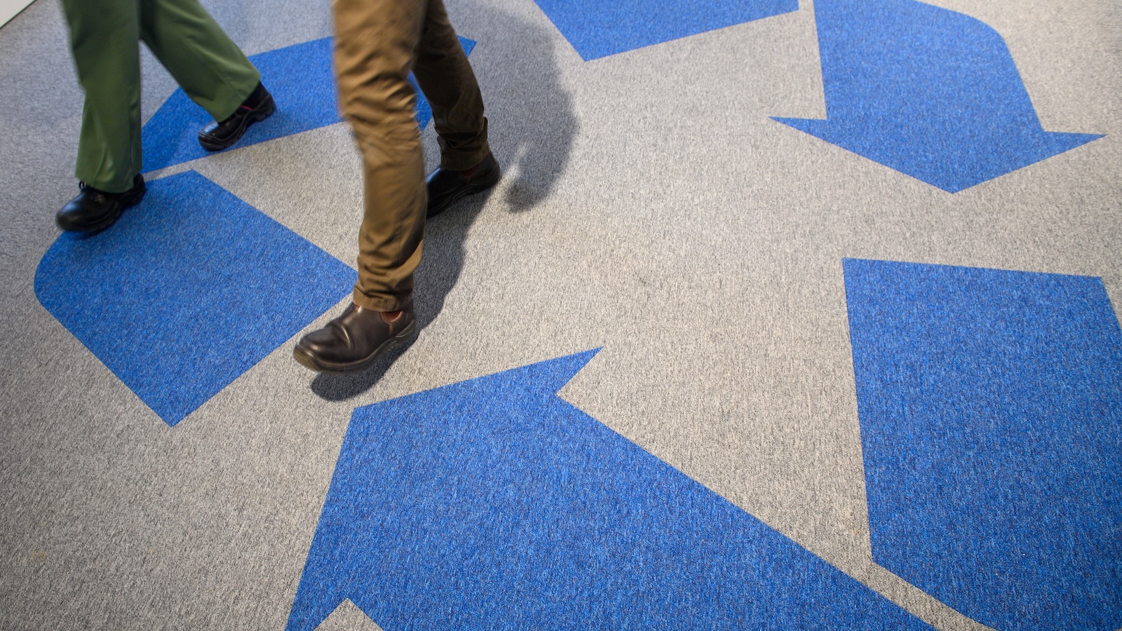 A large blue chasing arrows recycling symbol on the floor, with two people walking on top of it.