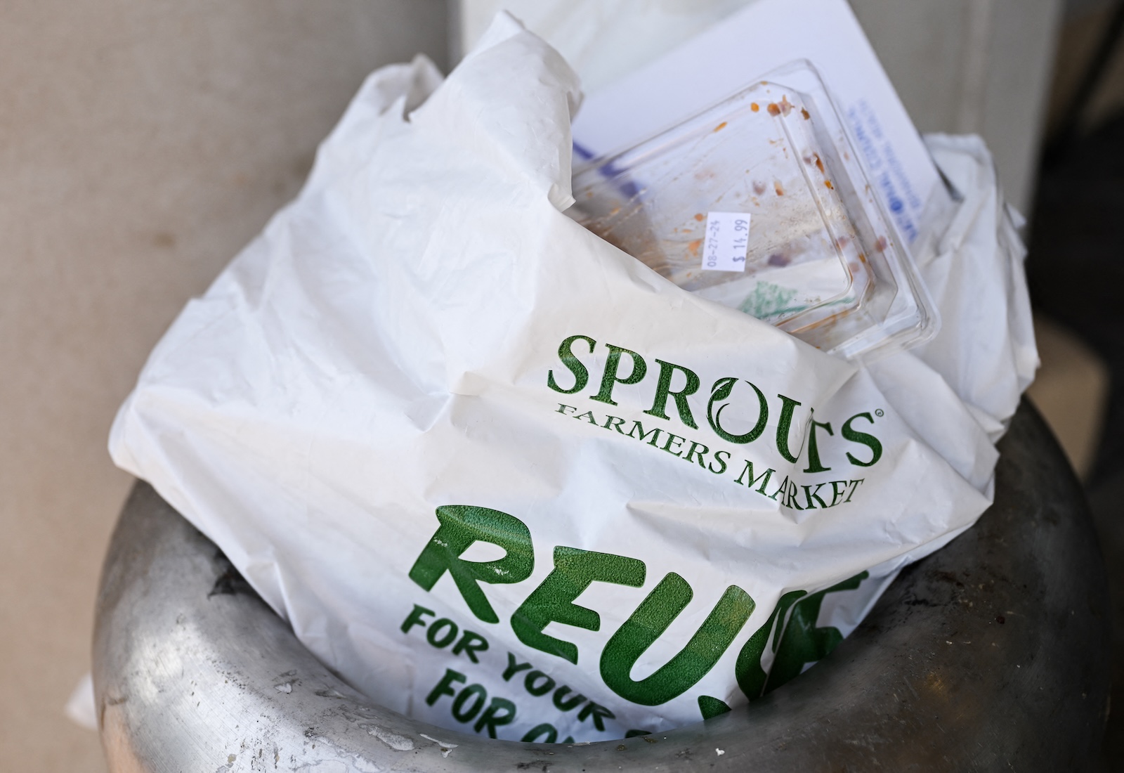 A thick plastic bag sits in a trash can. The word "Sprouts" is printed in green on the bag, and part of the word "reuse" is visible.