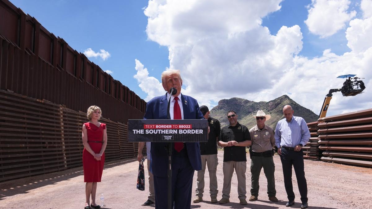 Donald Trump speaking behind a podium along the border wall in Arizona; the sky behind him takes up half the frame, and one woman dressed in red stands to his right, while a row men stand off to his left.