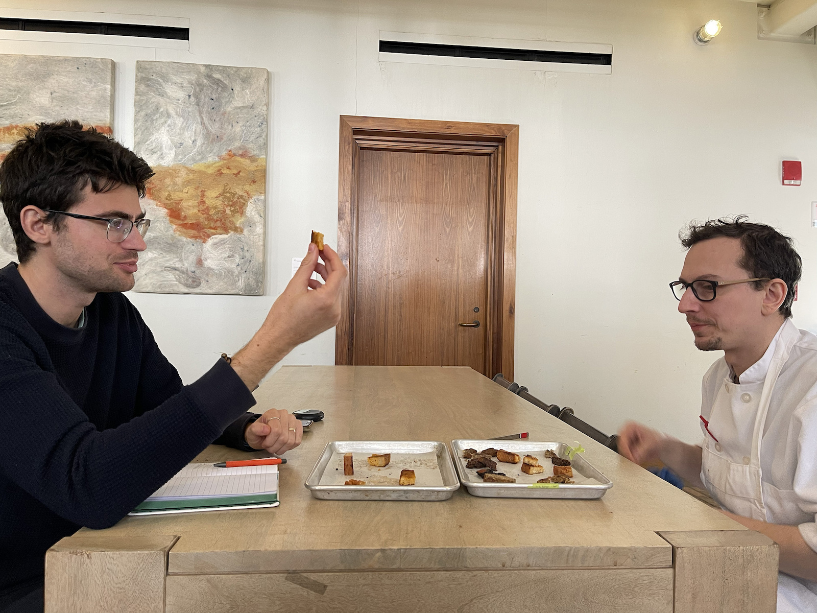 Two men sit at a table with food samples.