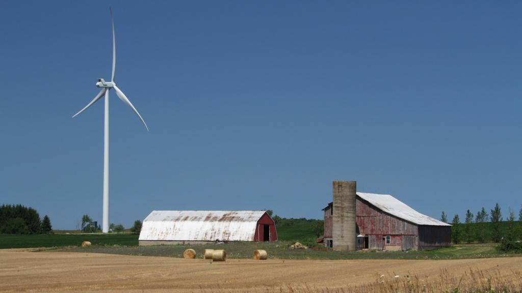 A windmill next to a barn with hay bales in front.