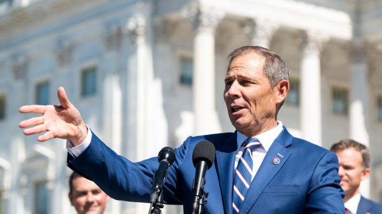 Photo of a man in a blue suit gesturing in front of a microphone in front of the Capitol