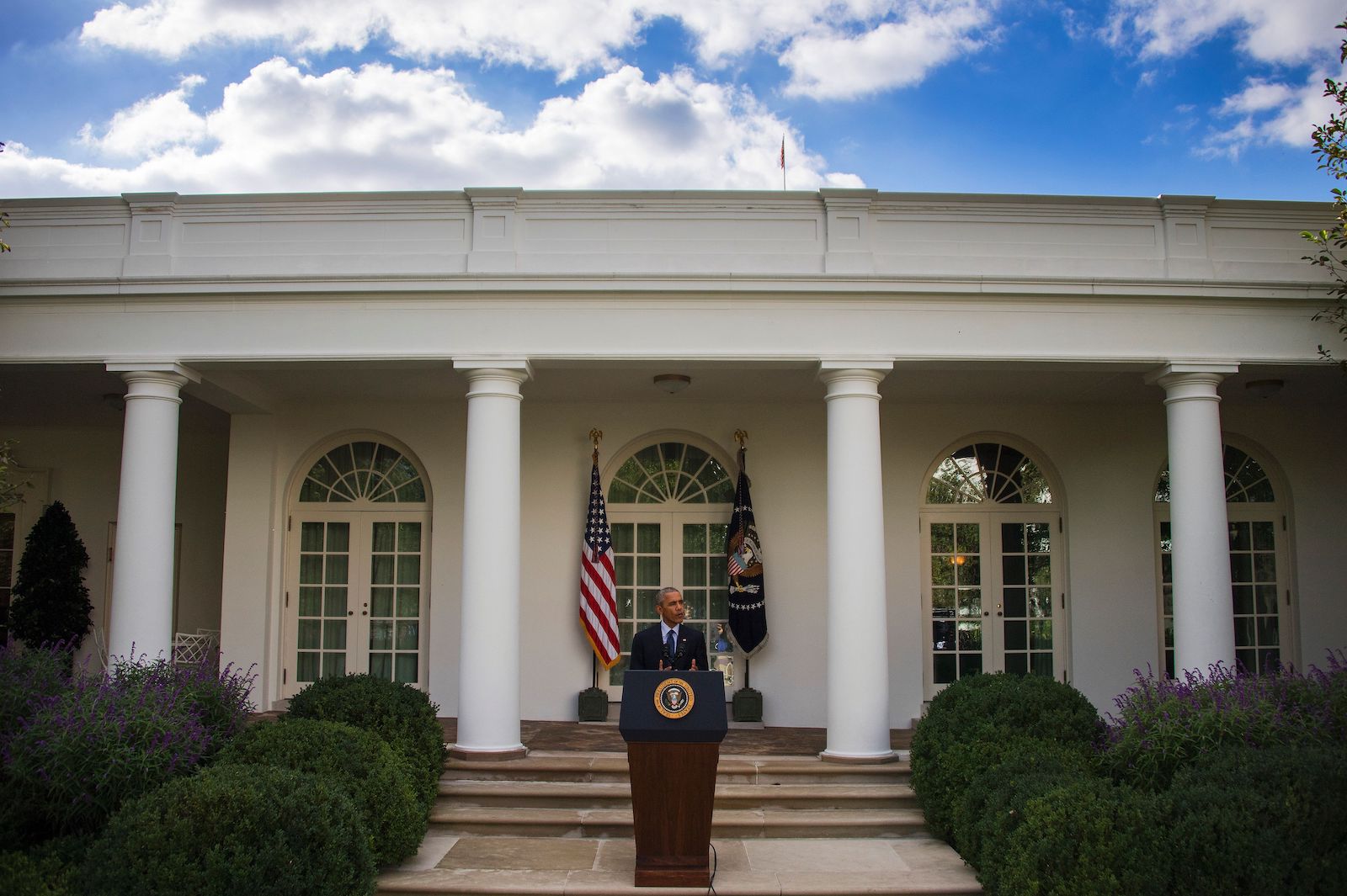 President Obama in a suit stands behind a podium in front of white pillars and stairs