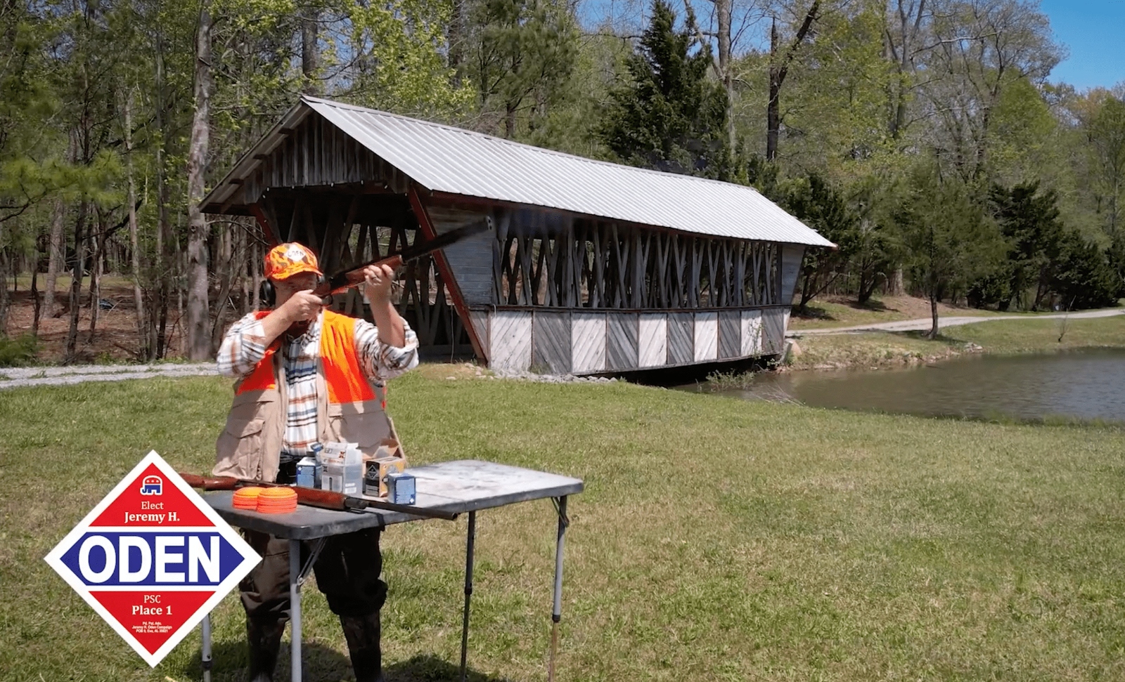 A man in an orange vest takes aim with a shotgun in a green field in front of a covered bridge