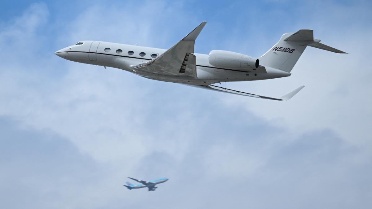 A small private jet flies through a blue sky with a passenger jet in the background