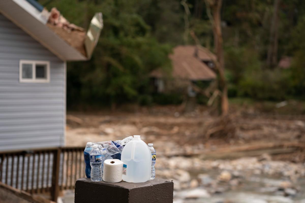 Bottled water and a toilet paper roll sit on a small box near a house