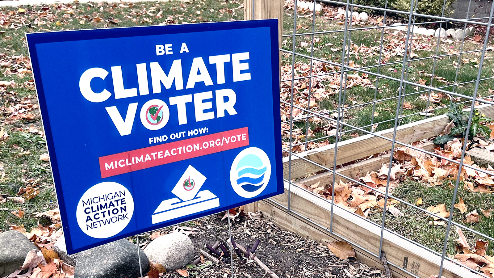 A blue sign on a green lawn covered in fall leaves says 