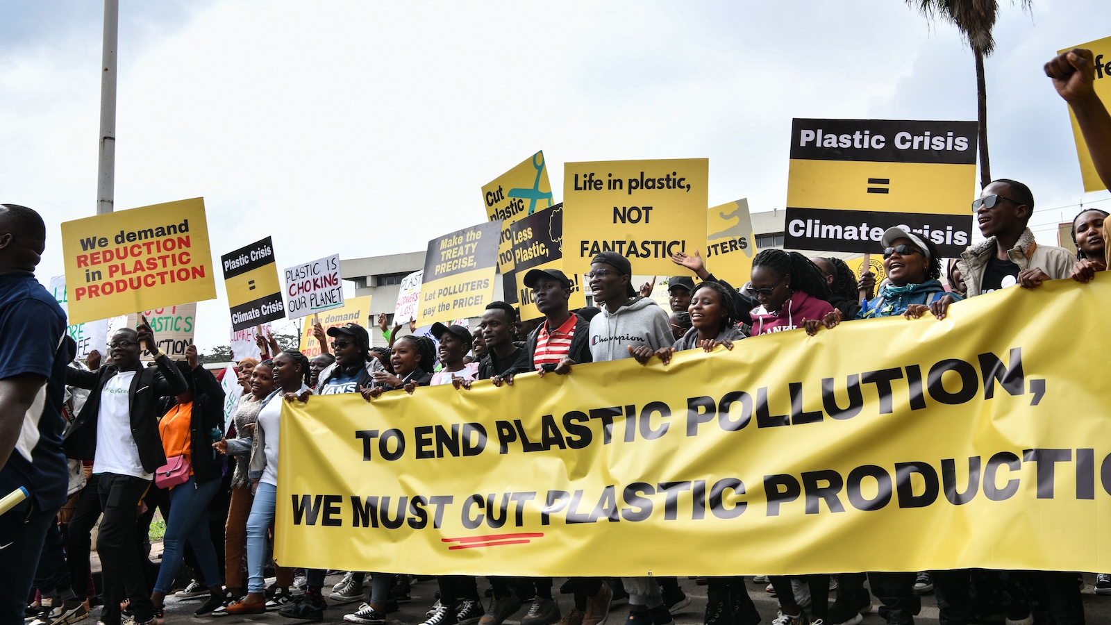 People hold up signs and a large horizontal banner reading "To end plastic pollution, we must cut plastic production"