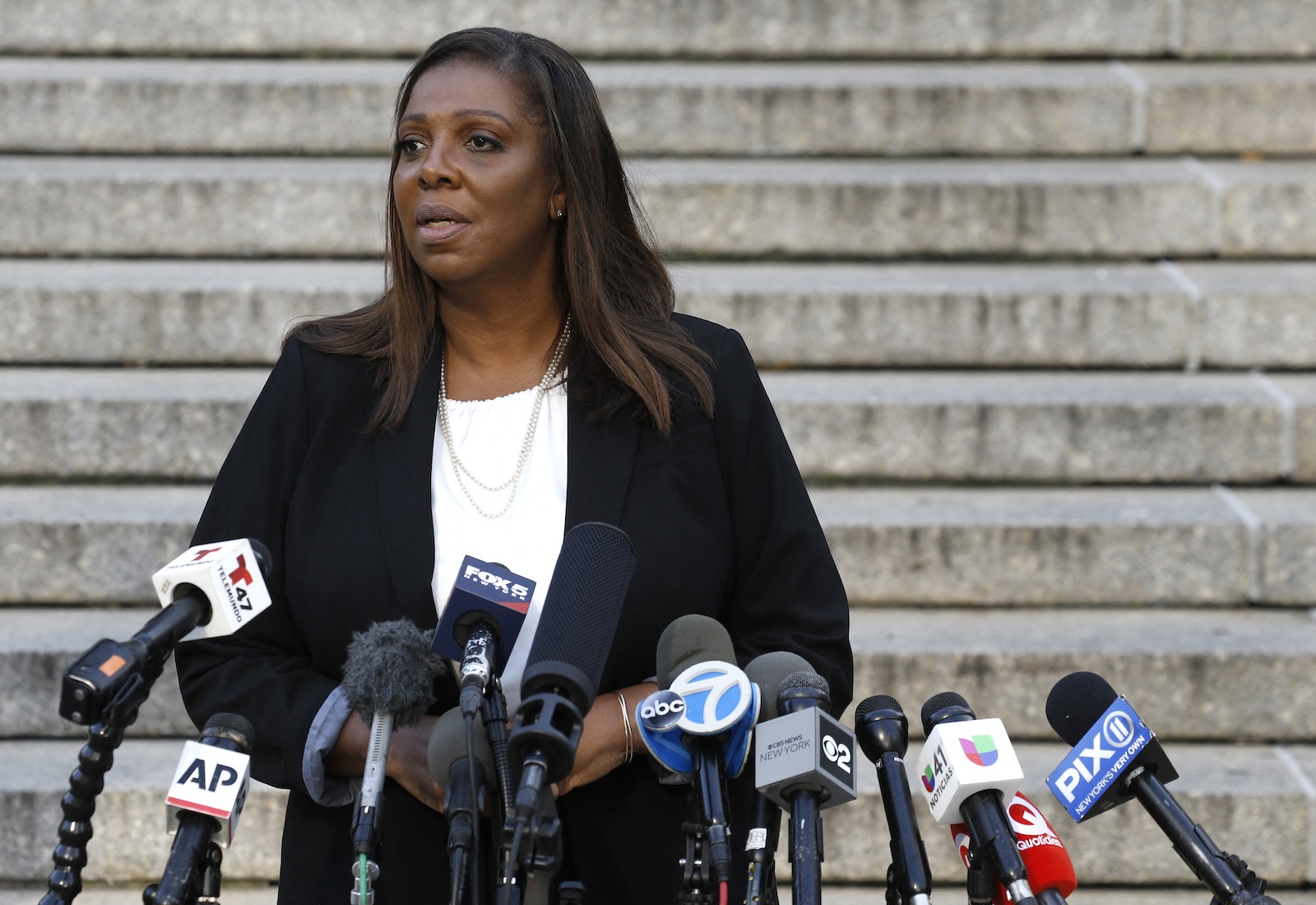 NY Attorney General Letitia James stands in front of stairs wearing a black jacket, with many news microphones pointing toward her.