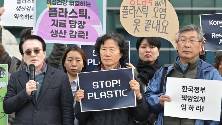 A small group of protesters hold signs, mostly in Korean, but one reads "Stop Plastic"