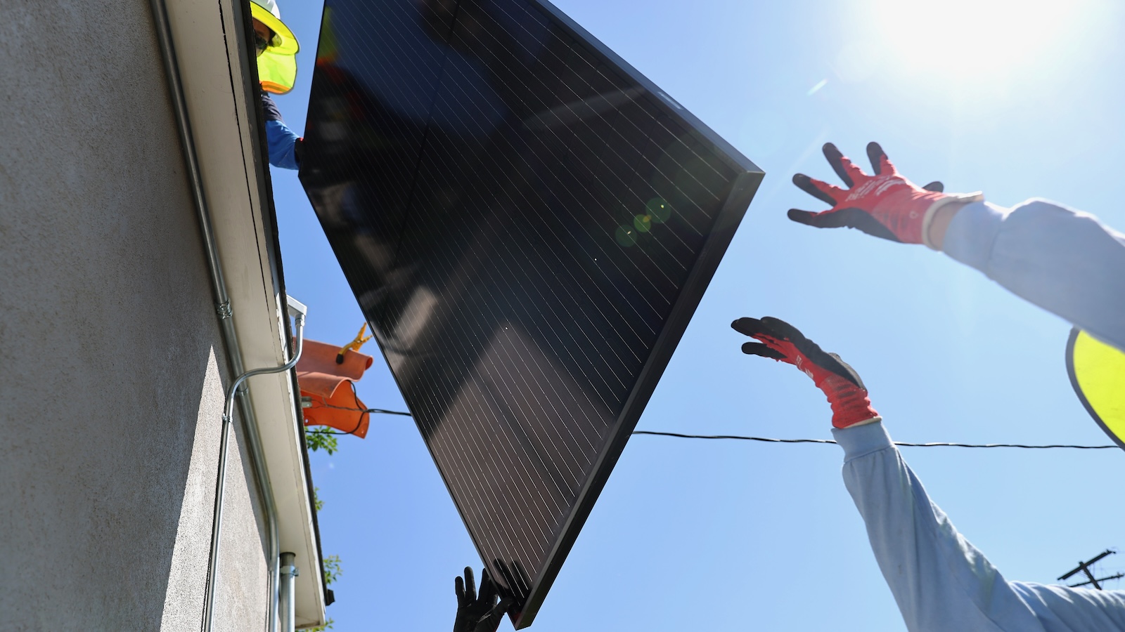 Two workers hand a solar panel to another worker on the roof a house.