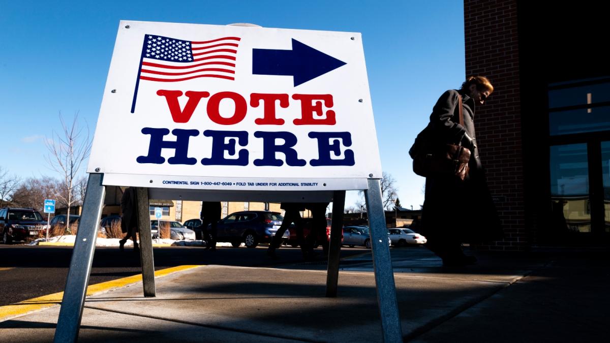 A large white vote here sign has a flag and arrow on it in front of a polling place