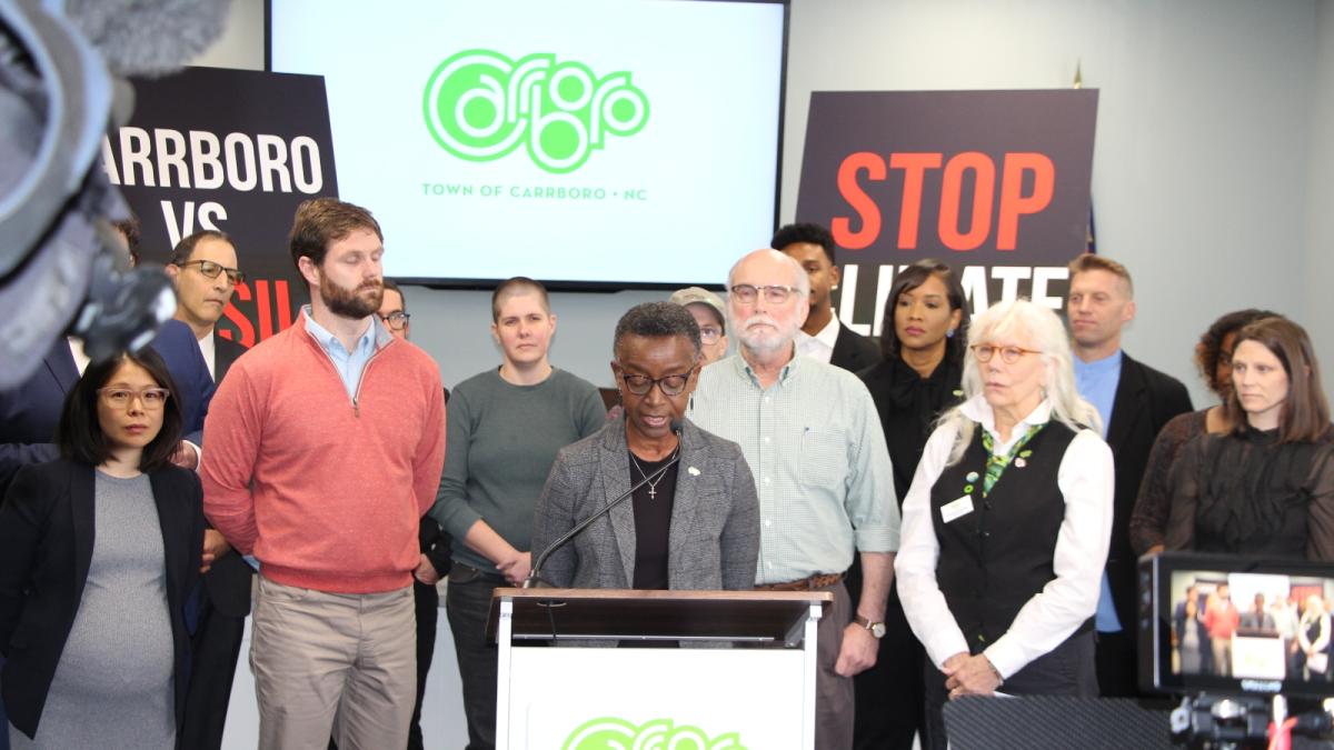 A woman in a gray jacket and black shirt stands at a podium with a group of people