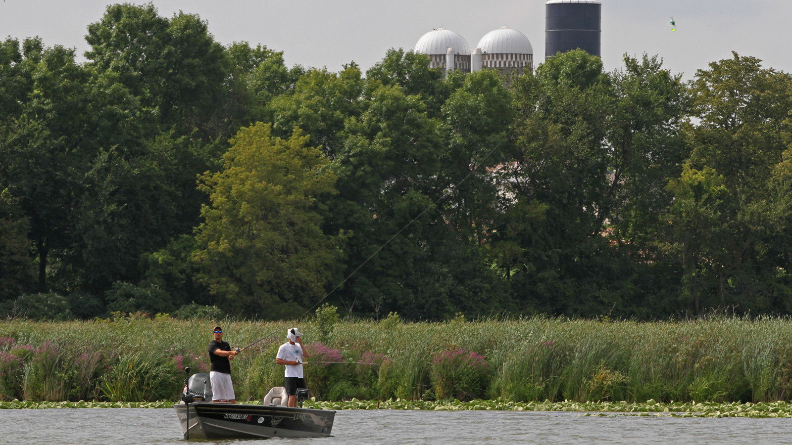 Two fishermen in a boat on a lake near a farm.