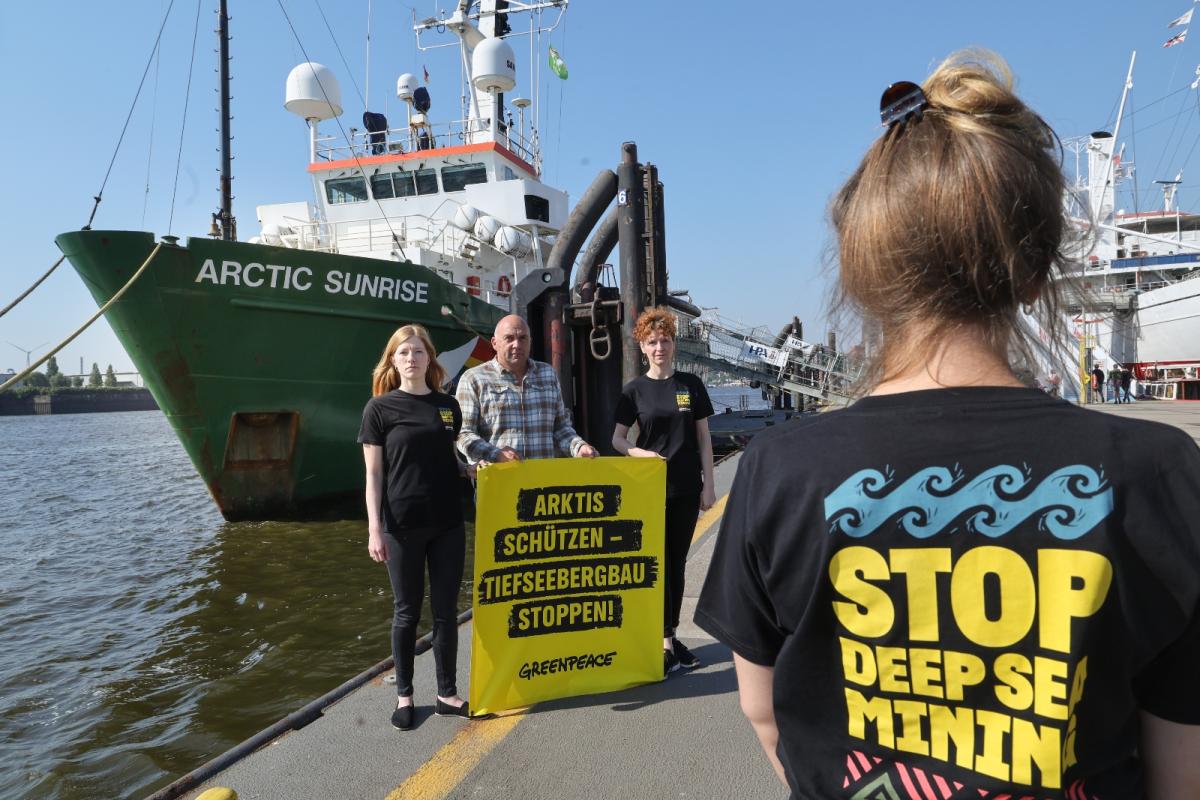 Activists from Greenpeace stand next to a ship called the Arctic Sunrise. In the foreground, an activist stands with her back to us, and her shirt reads Stop Deep Sea Mining.