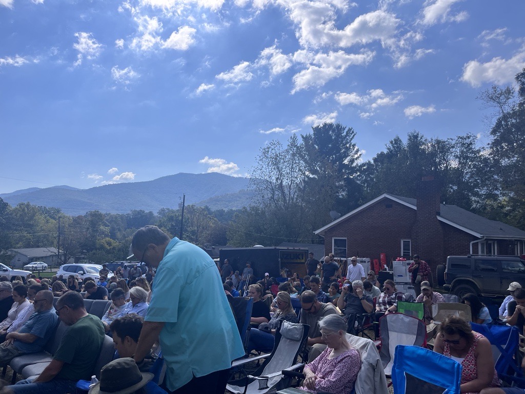 A group of people sit in folding chairs under a blue sky with mountains in the background. Their heads are bowed. A man in a blue shirt and glasses is standing with his head bowed.