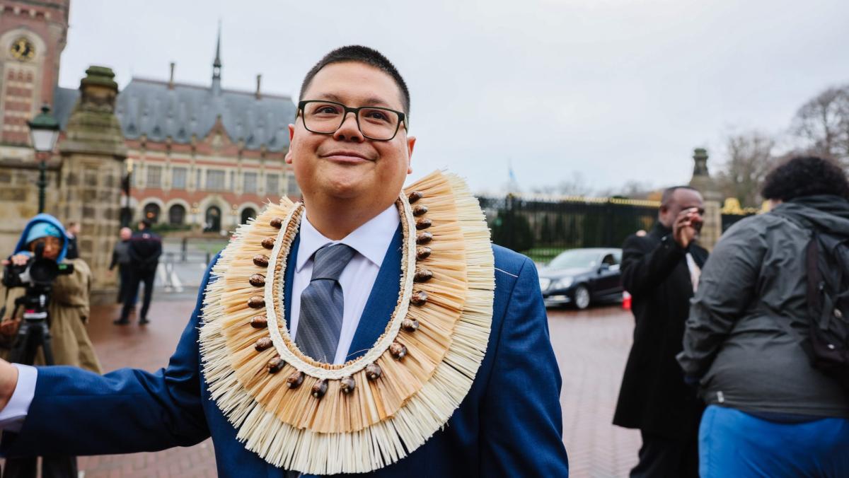 A man in a blue suit with shell necklace smiles in front of the Hague