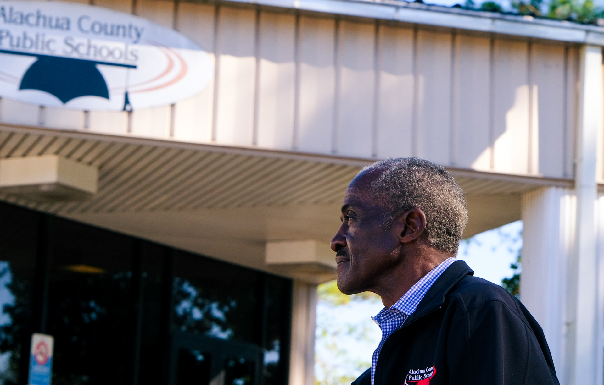 A man stands in profile in front of a building. His hair is cropped short and grey. The building has a sign that says, "Alachua County Public Schools."